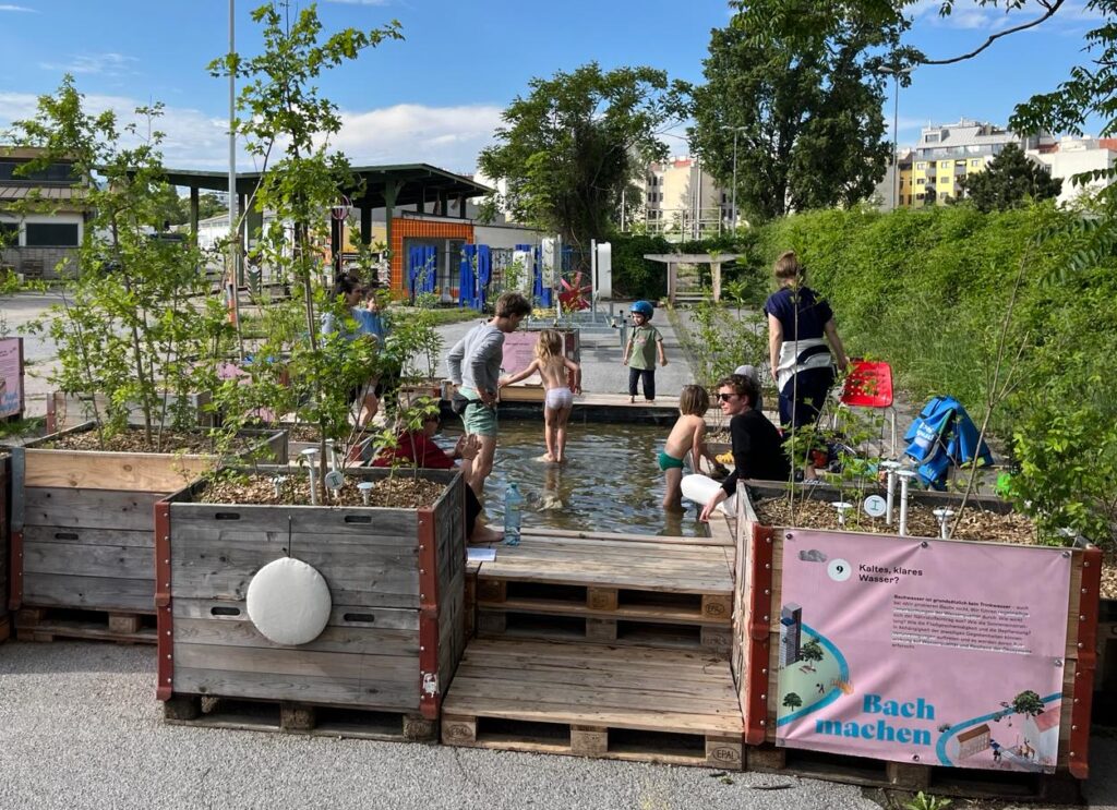 Ein Foto der temporären Bach-Installation bei der Klimabiennale. Man sieht Kinder im Wasser spielen und Erwachsene daneben sitzen. Im Wasser sind Steine und rundherum kleine Bäumchen und andere Pflanzen.